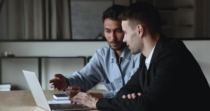 Positive busy diverse colleague men discussing project video presentation on laptop together, talking, pointing and looking at screen, cooperating on startup, brainstorming on ideas