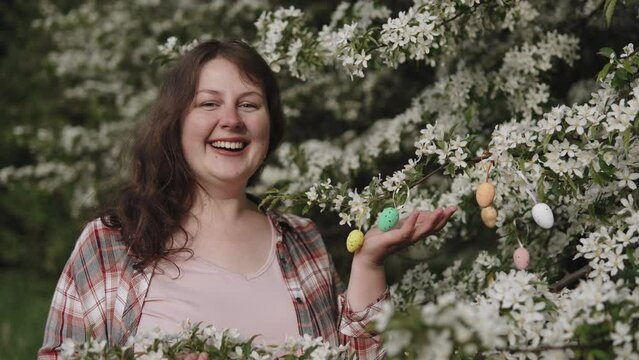 Portrait of simple happy plus size woman near flowering tree, push out shot. Female person smiles at the camera and touches with hand decorative colored easter eggs hanging on tree branch.