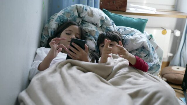 Children sharing cellphone screen in bed under bedsheets. Little brother and sister using modern technology device, engaged with entertainment media online, two kids watching content