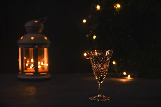 Glass with white wine with garland lights and candle lantern on the background. dark picture. Burning candle. Golden beverage in the glass. Beautiful lights reflected in the glass.