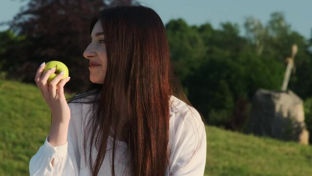 Young lady catches green apple against natural surrounding. Young woman eats apple while enjoying sun rays sitting in meadow