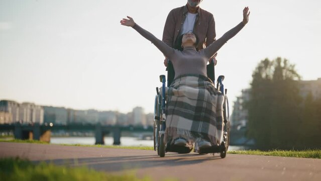 Elderly husband walking with wife in wheelchair. Senior family couple spending time in city park. Man caring woman showing love, they look happiness. Living with physical disability concept.