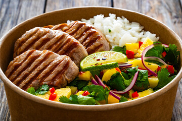 Grilled pork loin steaks with rice and mango salad in lunch box to go on wooden table
