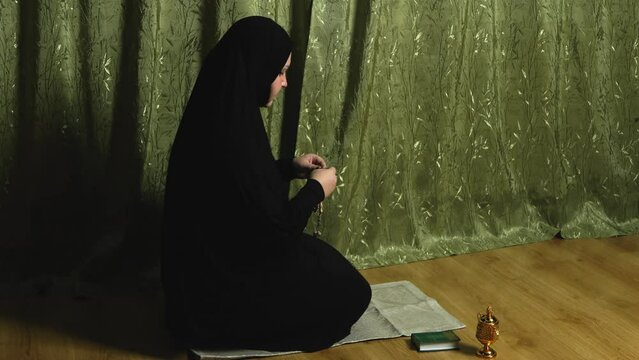 A Muslim woman in a black khimar in profile at home reads prayer while fingering her rosary on the rug. Overall plan