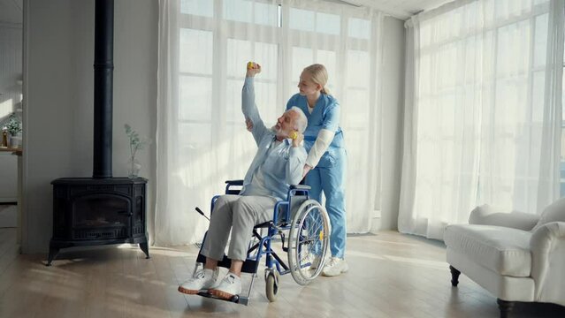 Nurse woman cares old man in wheelchair. Paralyzed elderly man does exercises under supervision of a nurse supporting at clinic. Living with spine legs paralysis, special needs people health concept.