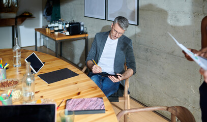 handsome man with gray hair wearing eyeglasses sitting at table with laptop and tablet and making notes on paper. Portrait of company executive writing report. Concept of business
