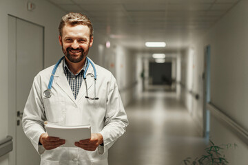 Male doctor in white gown with stethoscope standing in hall of clinic and holding documents in hands
