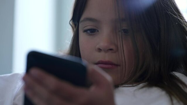 Close-up face of little girl holding and using phone with solemn bored expression. Child watching content online