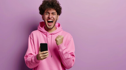 excited young man with curly hair in a pink hoodie, holding a smartphone with one hand and making a fist with the other