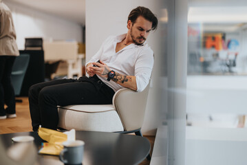 A pensive young man with tattoos sits in a stylish office lounge, looking at his smart watch, embodying modern business casual style.