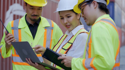 Three workers wearing hard hats are using tablets working together in a container yard, a shipping import and export business. Operating a logistics shipping yard business teamwork concept