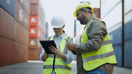 Two workers wearing hard hats are using tablets working together in a container yard, a shipping...