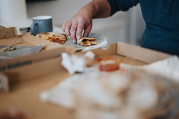 Close-up image of a person picking up a slice of pizza from a cardboard box, with a casual home environment in the background.