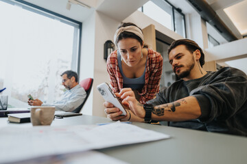 Two engaged young professionals discuss work on a smart phone in an office setting, showcasing teamwork and collaboration.