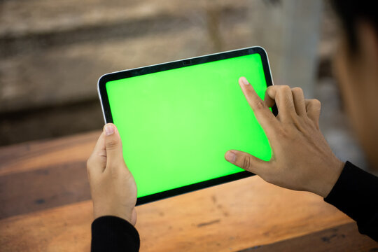 Mock up photo of a close up shot featuring a mans hand holding an iPad tablet with a green screen against the background of a wood cafe table