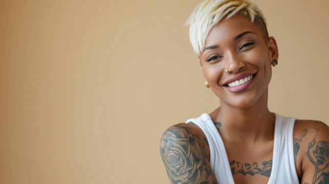 smiling woman with blonde bob hair, a nose piercing, and visible tattoos on her shoulders is wearing a white tank top and looking at the camera