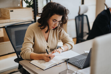 A businesswoman is deeply focused while writing notes at her workstation. She's surrounded by a...