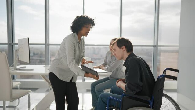 Paralyzed man works with colleagues. Guy in wheelchair has spine trauma discussing working project in office on meeting with diversity team. Teamwork brainstorming, special possibilities needs.