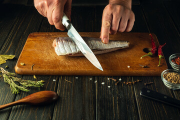 Hands of a chef using a knife to cut fresh Hake fish on a wooden cutting board. Home-cooked national fish dish according to a unique recipe