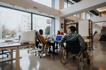 Modern office setting showcasing diversity with a man in a wheelchair and multiethnic colleagues working. - obrazy, fototapety, plakaty