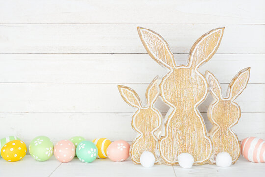 Rustic style Easter decor on a white shelf against a white wood panel background. Wooden bunnies and pastel Easter eggs.