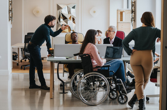 A cross generational group of professionals engage in a business meeting. The image includes a person using a wheelchair, depicting inclusivity and teamwork in a modern office setting.