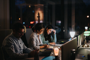 Concentrated business colleagues of various ages working intently at their desks during a late evening in a modern office space, illuminated by desk lamps.