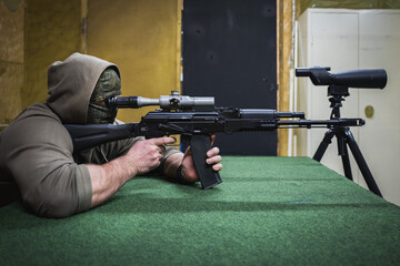 A male shooter in a mask and hood shoots with an AK 74m with an optical sight at a shooting range.