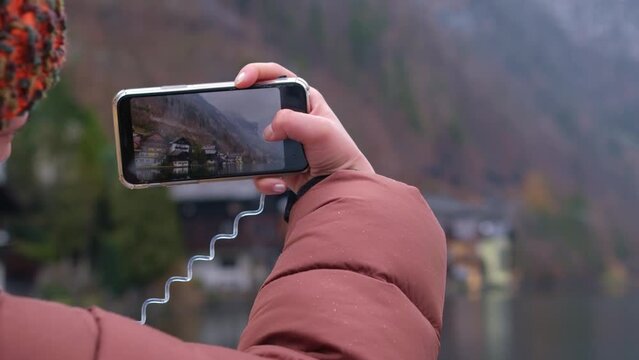 Hand with a phone records a video of a beautiful landscape. Tourist takes a photo of the village of Hallstatt. View of Hallstatt, Austria. Boat passenger records the view of the shore from the water.
