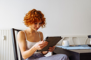 red-haired teenage girl sits in dining room at table, uses tablet, chats