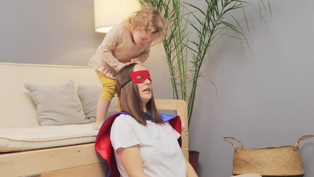 Happy cheerful blonde wavy haired infant girl trying to sit on mother shoulders enjoying funny playing at home on floor in living room funny active family games