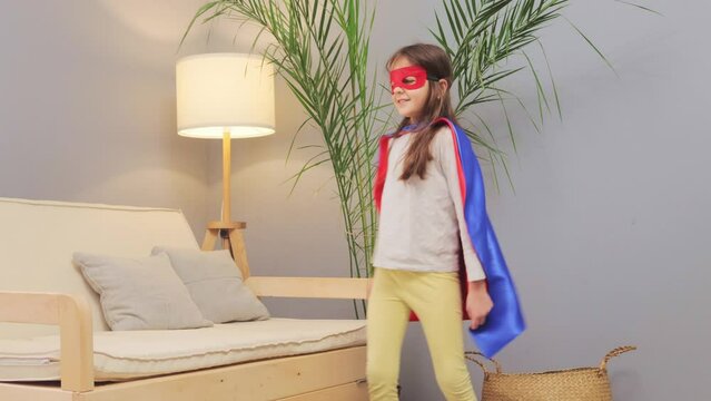 Overjoyed playful little brown haired girl wearing superhero costume and mask playing at home in living room jumping and dancing displaying leadership and joy