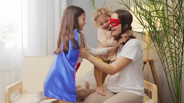 Cheerful woman wearing superhero mask playing with her daughters at home sitting on sofa in living room hugging raising fist like flying to save work laughing happily