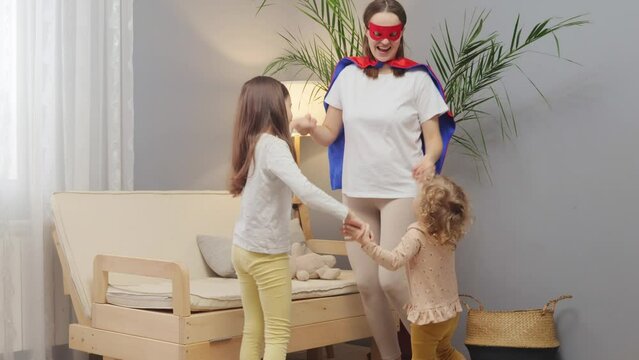 Cute kids playing together with their mother at home wearing superhero costumes dancing and jumping while holding hands enjoying active role play in living room