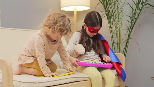 Playful active little sisters kids playing dressing up superhero together in living room at home play with toy piano and interactive tablet sitting on sofa in home interior
