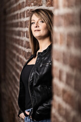 Woman model around thirty in black leather jacket and hot pants in front of urban brick wall.