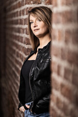Woman model around thirty in black leather jacket and hot pants in front of urban brick wall.