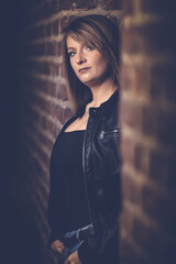Woman model around thirty in black leather jacket and hot pants in front of urban brick wall.