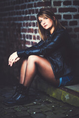 Woman model around thirty in black leather jacket and hot pants in front of urban brick wall.