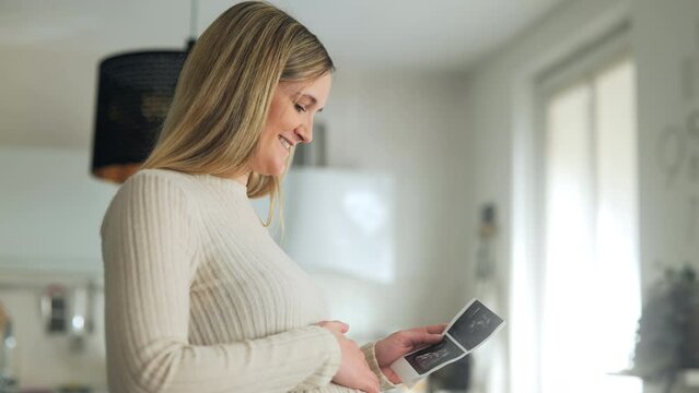 Maternity love. Beautiful and happy pregnant woman admiring sonography picture of her unborn baby and caressing belly, standing at home, slow motion