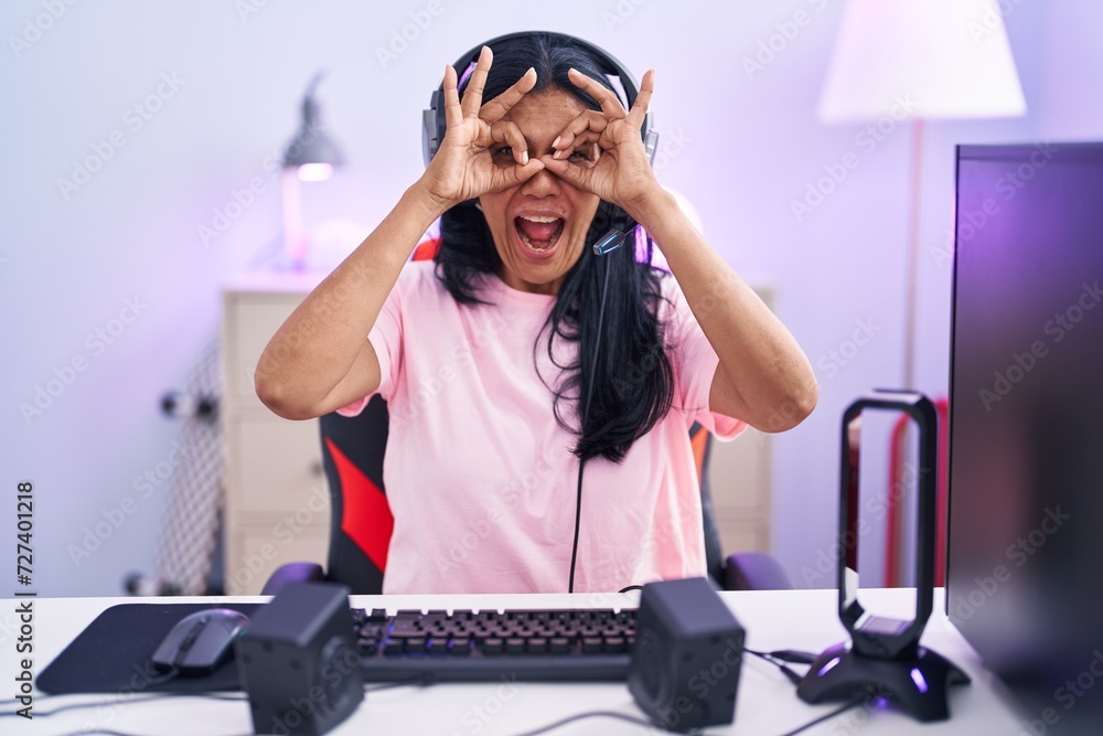 Poster Mature hispanic woman playing video games at home doing ok gesture like binoculars sticking tongue out, eyes looking through fingers. crazy expression.