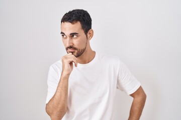 Handsome hispanic man standing over white background with hand on chin thinking about question, pensive expression. smiling with thoughtful face. doubt concept.