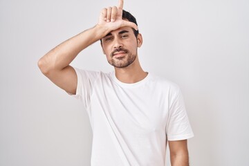 Handsome hispanic man standing over white background making fun of people with fingers on forehead...