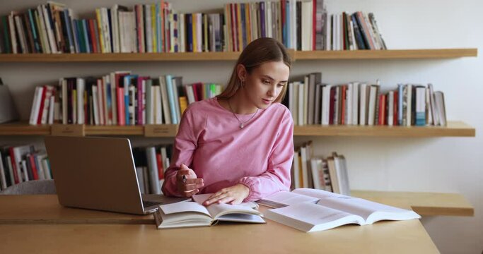 Pretty focused European student girl sit at library desk, makes notes in copybook, preparing for exams, work on project, read topics in textbook, learn theory, gain new knowledge. Education, studying