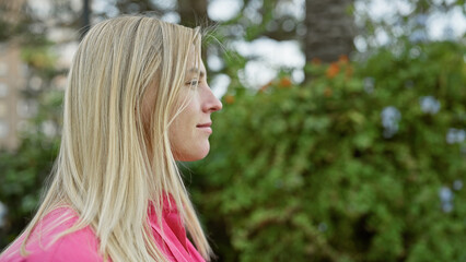 Profile of a thoughtful blonde woman in a pink jacket outdoors by greenery in a park setting.