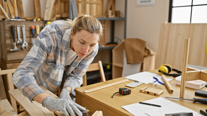 A focused young caucasian woman sands wood in a well-equipped indoor carpentry workshop.