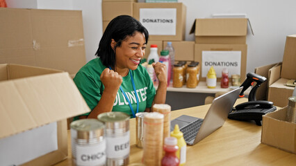 Excited hispanic woman volunteering at a food donation center surrounded by boxes and goods.