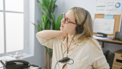 A pained young woman in headphones touching her neck in modern office setting, showing signs of...