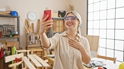 Smiling woman takes a selfie in a workshop with woodwork tools on a workbench behind her.