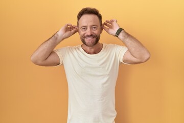 Middle age man with beard standing over yellow background smiling pulling ears with fingers, funny gesture. audition problem